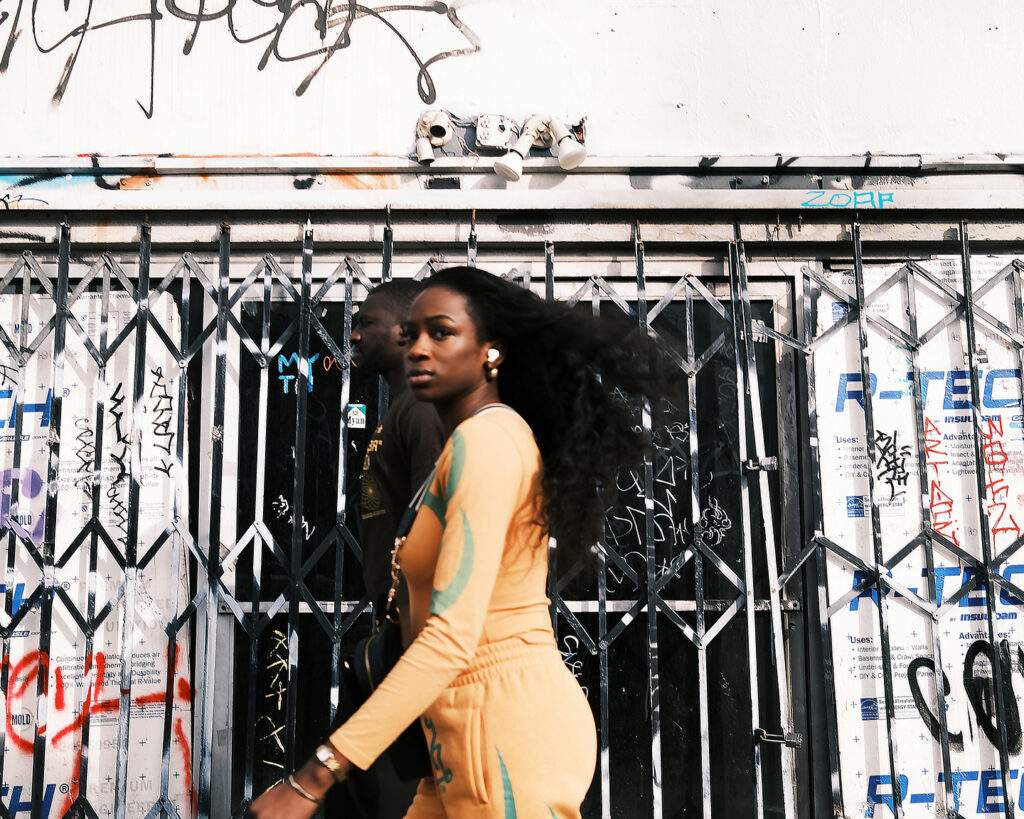 Xan Walker, What Are You Looking At?, a woman in yellow glances at the camera I front of a security gate in the Mission, 2026, Archival pigment print, 8" x 10"
